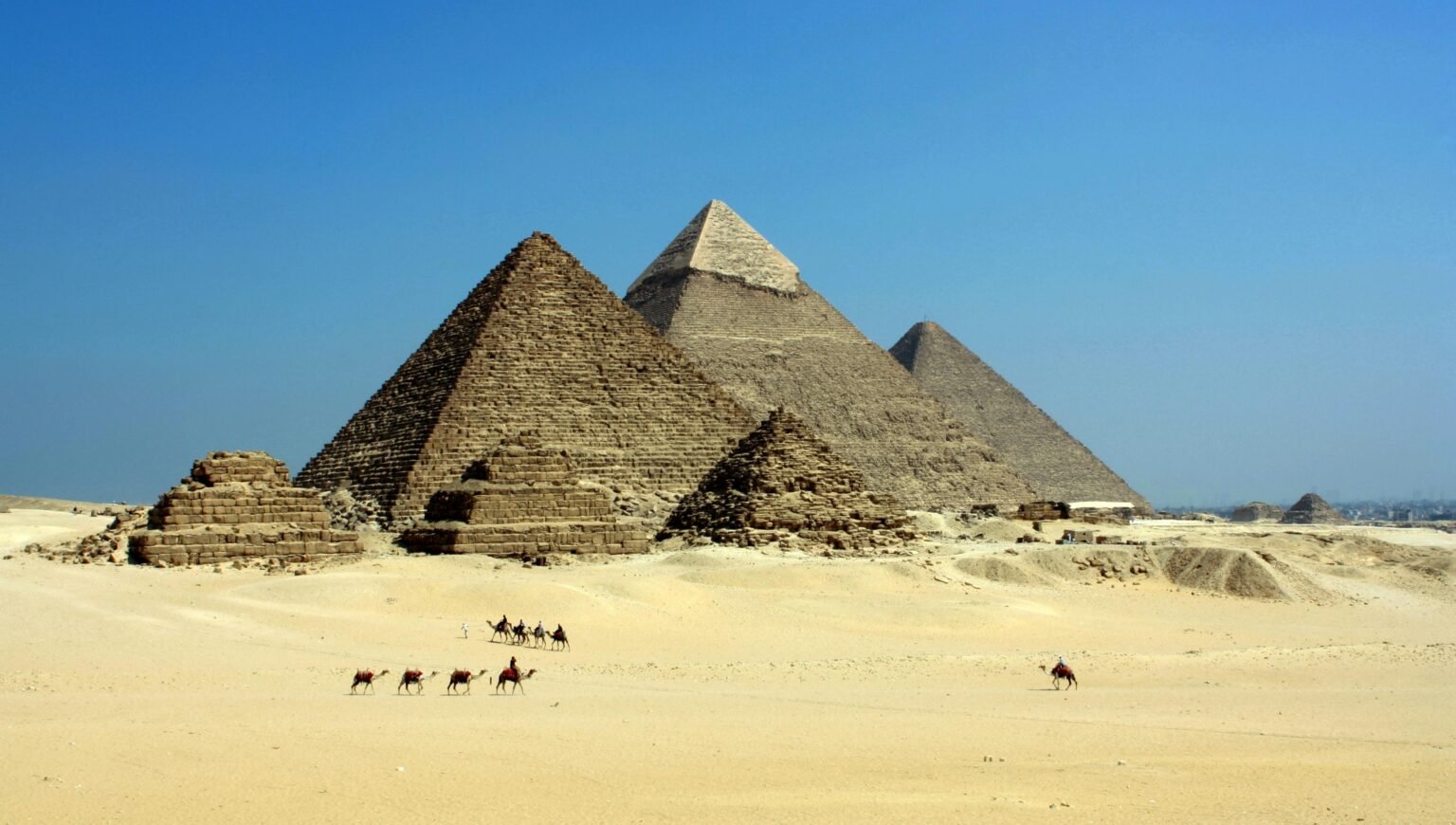 Capture of the iconic Pyramids of Giza under a clear blue sky with camels traversing the sandy desert.